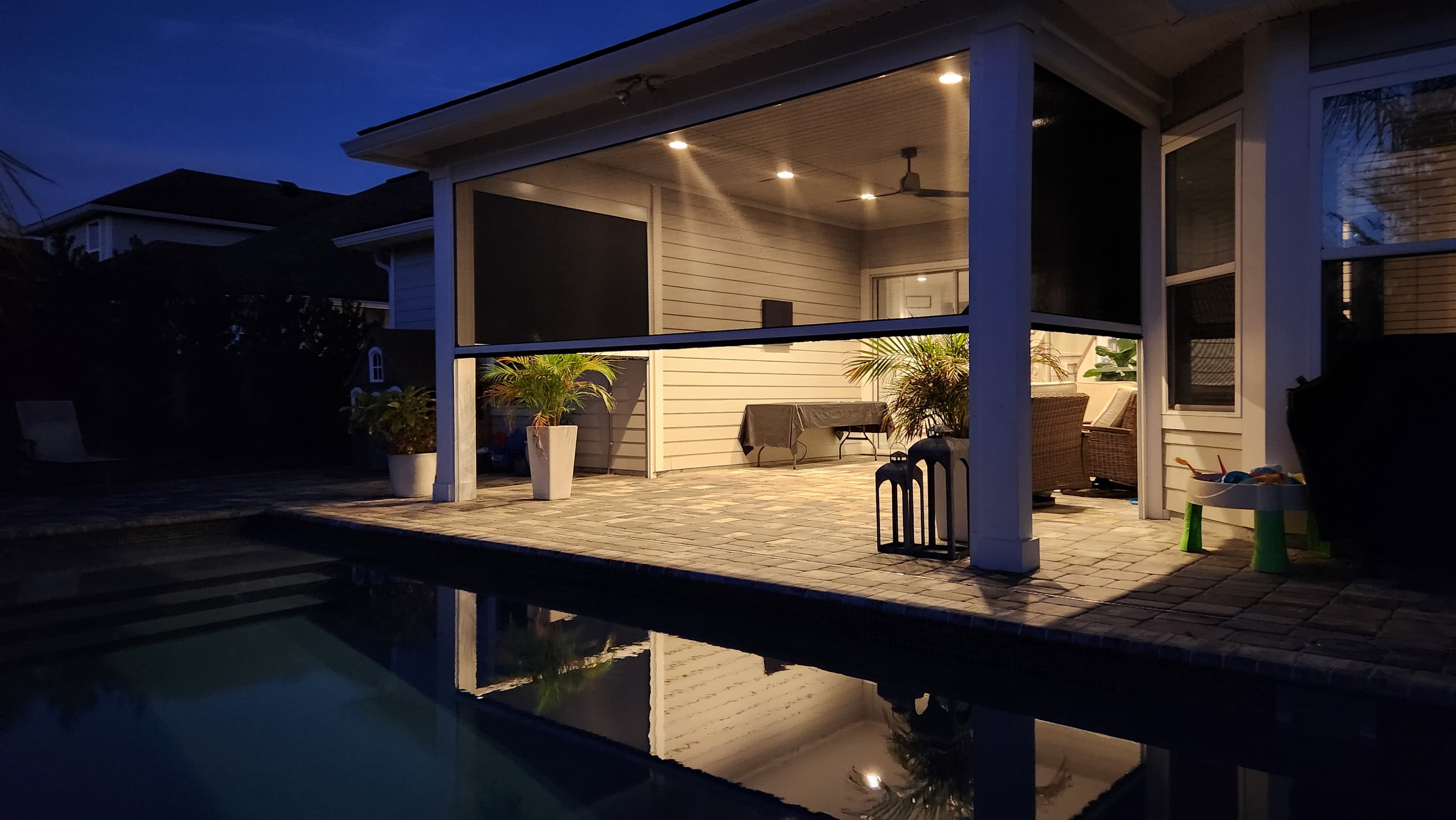 Motorized insect screens enclosing a warmly lit covered patio beside a pool at dusk